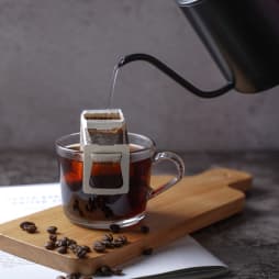Повна чашка чаю на дерев'яній дошці, в яку доливають окріп з чайнику